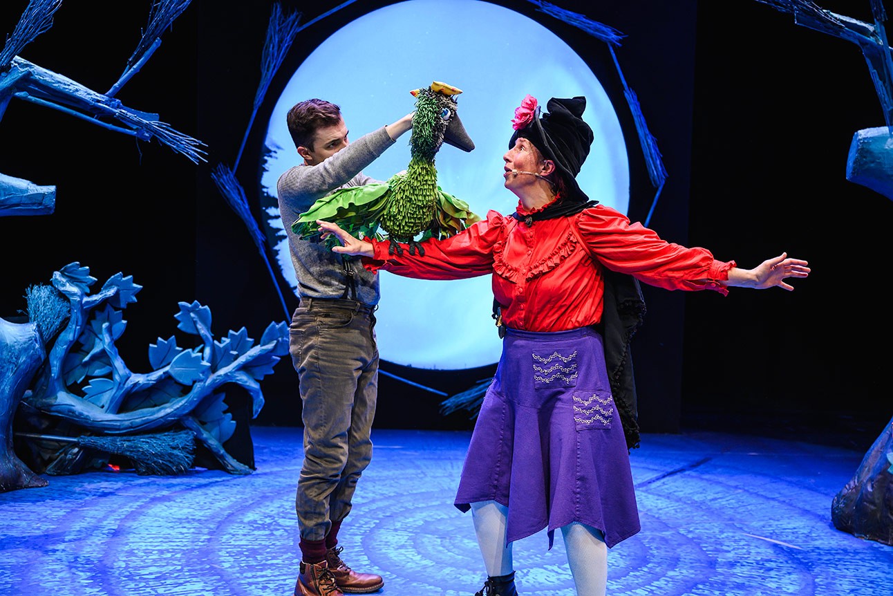 Two performers on stage; one holds a green bird puppet, the other wears a witch costume.