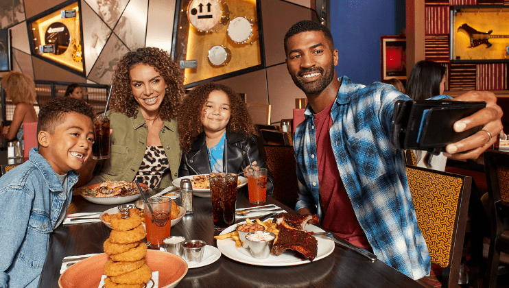 Family of four smiling at a the Hard Rock Cafe, with ribs, onion rings, and drinks.