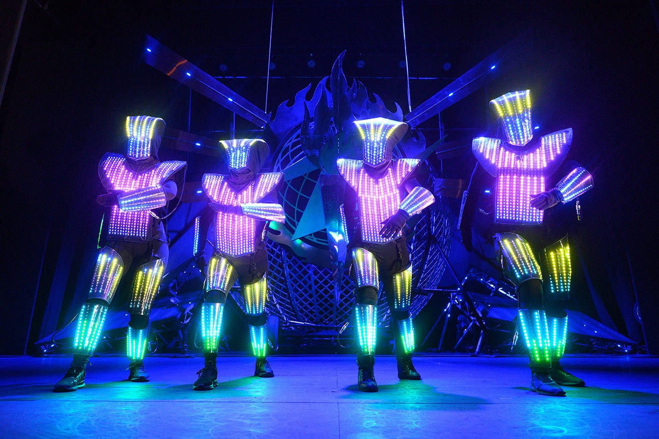 Four performers in illuminated costumes with neon stripes, posing on stage against a dark background.