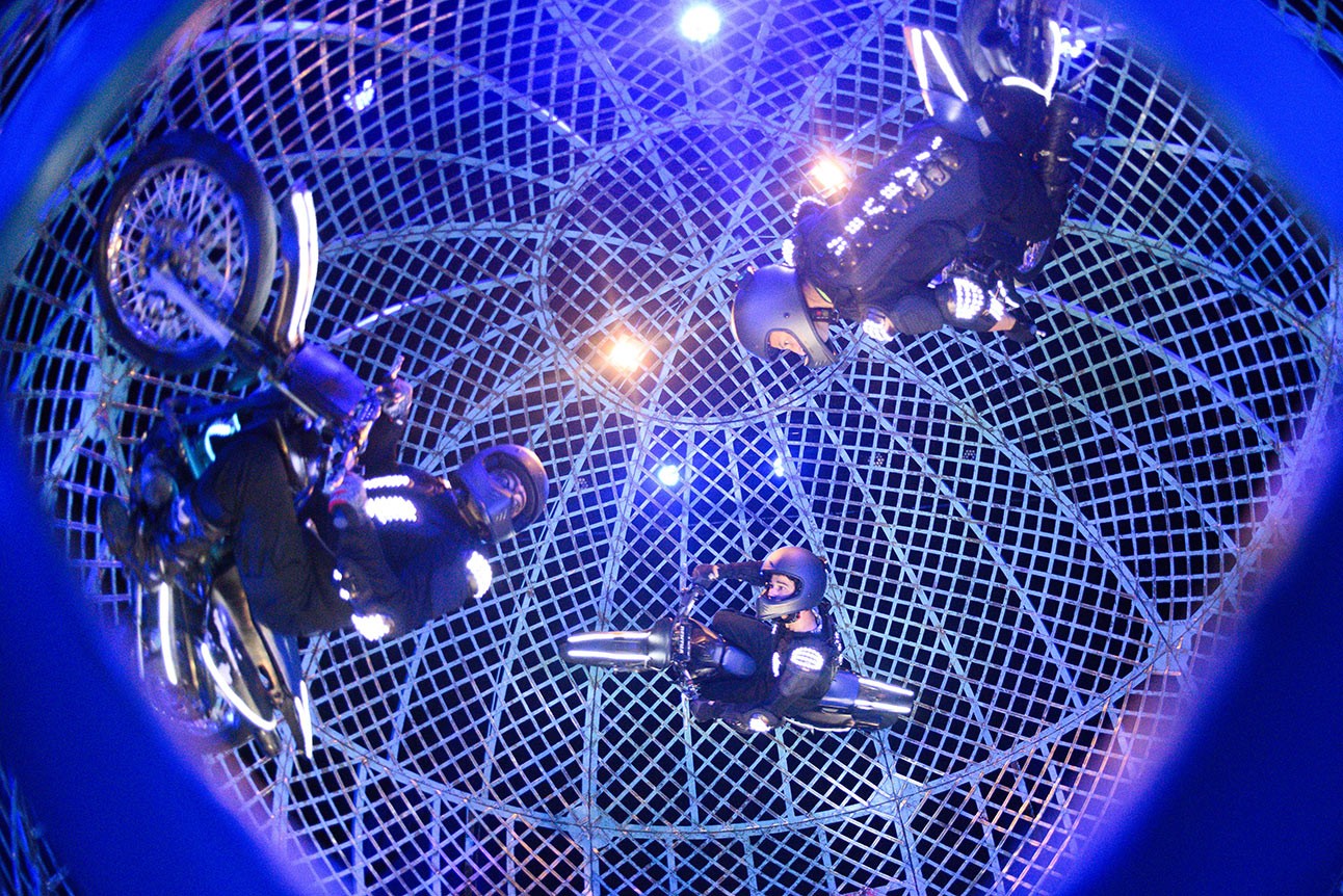 Three motorcyclists perform inside a spherical metal cage, illuminated by blue and purple lights.