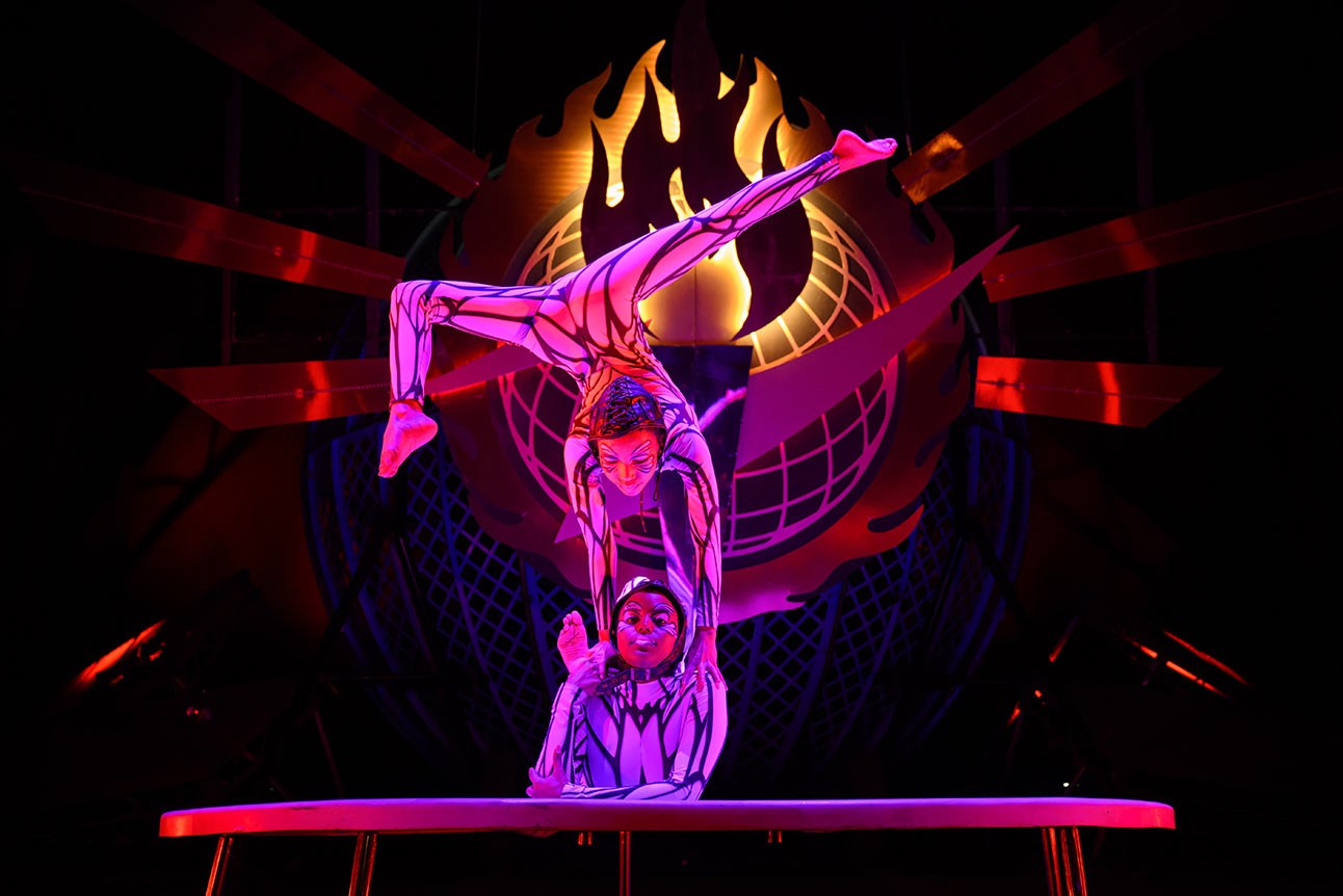 Two acrobats perform a balancing act on stage, with vibrant purple lighting and fiery backdrop.