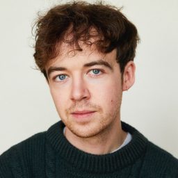 A person with curly brown hair and a dark jumper, looking directly at the camera.