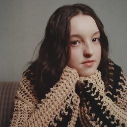 Person with long hair wearing a beige and black knitted sweater, resting chin on hands, neutral background.