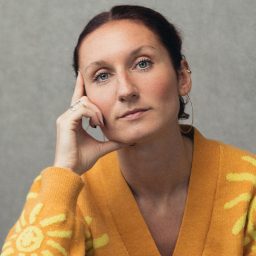Woman with brown hair in orange sweater, resting chin on hand, grey background.