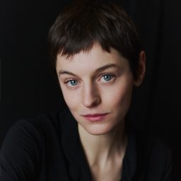 A person with short brown hair and blue eyes, wearing a black top, against a dark background.