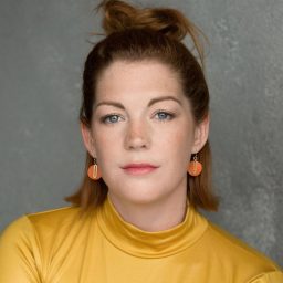 Woman with red hair in yellow top, orange earrings, against grey textured background.