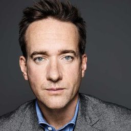 A man with short brown hair in a grey suit jacket against a dark grey background.