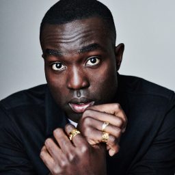 Paapa Essiedu headshot; he rests chin on hands, wearing black jacket, gold rings, neutral background.