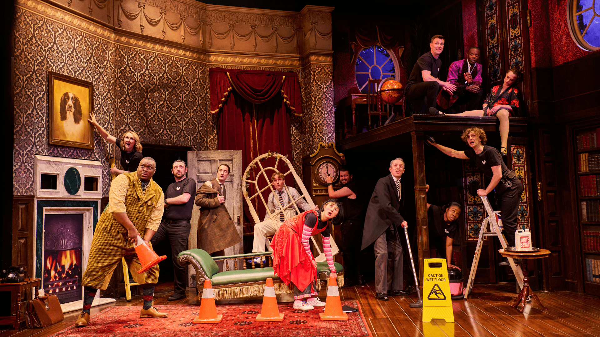 The Play That Goes Wrong Actors in a chaotic stage scene with vintage decor, props, and a "wet floor" sign.