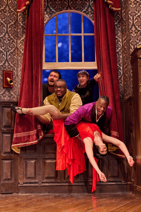 Four actors in colourful costumes comically struggle through a window, holding a woman in a red dress.