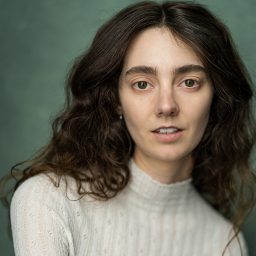 Woman with long curly hair, wearing a white top, against a green background.