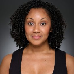A woman with curly hair and a black top against a dark grey background, looking forward.