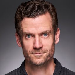 A man with short curly hair and a serious expression against a grey gradient background.