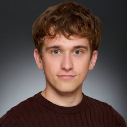 Young man with short brown hair wearing a dark jumper, neutral expression, grey gradient background.