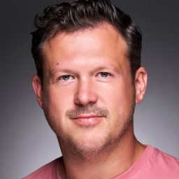 A person with short hair wearing a pink shirt against a grey background, looking forward.