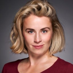 A woman with short blonde hair, wearing a burgundy top, smiles slightly against a grey background.
