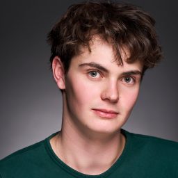 Young man with short brown hair wearing a green shirt, neutral expression, grey background.