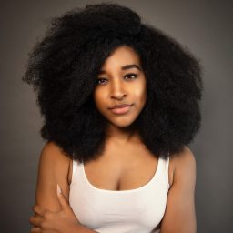 A person with voluminous curly hair wearing a white vest top against a plain grey background.