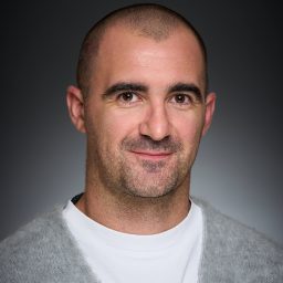 A man with a shaved head and grey sweater smiles slightly against a dark grey background.
