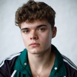 Young man with curly hair in a green and black jacket, neutral expression, plain background.