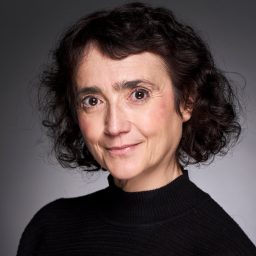 A woman with short, curly dark hair wearing a black turtleneck against a grey background.