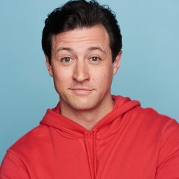 A man in a red hoodie poses against a light blue background, looking directly at the camera.