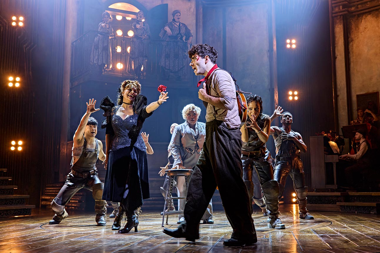 Performers on stage in "Hadestown," dramatic lighting, one holding a red flower, others gesturing.