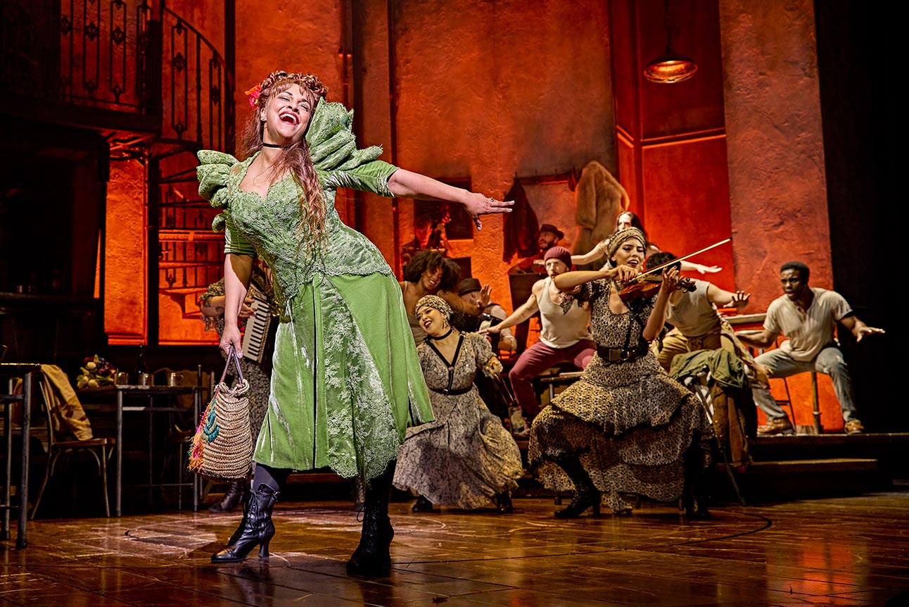 Danielle de Niese performs energetically in a green dress with the cast of Hadestown on stage.