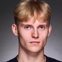 Young person with short blond hair, neutral expression, wearing a dark top against a grey background.
