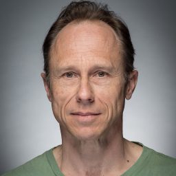 A man with short brown hair wearing a green shirt, neutral expression, grey background.