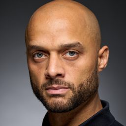 A man with a shaved head and beard, wearing a dark shirt, against a grey background.