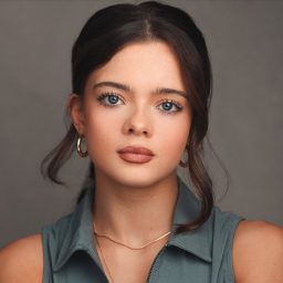 Young woman with dark hair, wearing a sleeveless top and earrings, against a grey background.