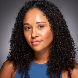 A woman with curly hair, wearing a blue top, poses against a grey background.