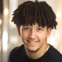 Smiling man with curly hair and earrings, wearing a black shirt, blurred light background.