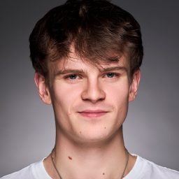 A young man with brown hair wearing a white shirt, neutral expression, grey background.