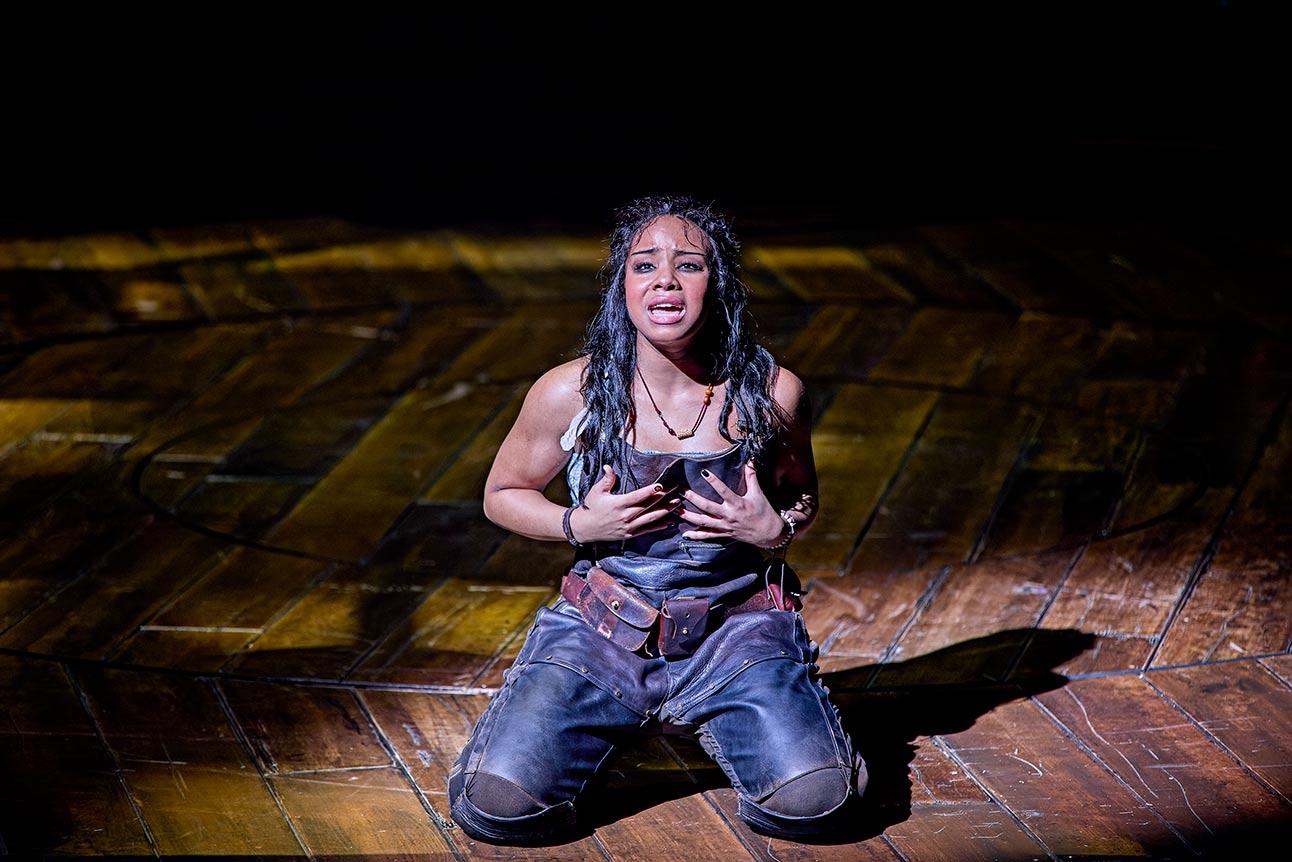 Fayth Ifil kneels on stage, wearing dark overalls, expressive and emotional, under dramatic lighting.