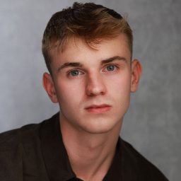 Young man with short hair, wearing a dark shirt, poses against a grey studio background.