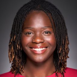 Smiling person with shoulder-length braided hair, wearing a red top, against a grey background.