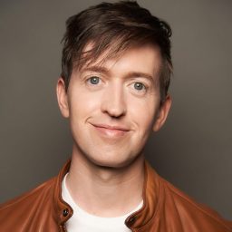 Smiling man with short brown hair, wearing a brown jacket against a plain grey background.