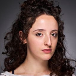 A woman with curly dark hair and a neutral expression against a grey background.