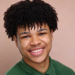 Smiling person with curly hair wearing a green shirt against a plain beige background.