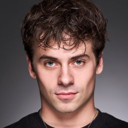 A man with short, dark hair and a neutral expression against a grey background.