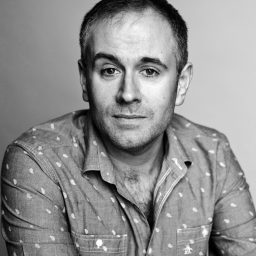 A man with short hair wearing a patterned shirt, looking directly at the camera, neutral background.