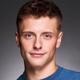 A person with short hair wearing a blue shirt, smiling slightly against a grey background.