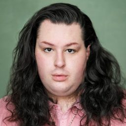 Person with long dark hair, wearing a pink shirt, against a plain green background.
