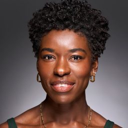 Smiling woman with short curly hair, wearing earrings and a necklace, against a grey background.