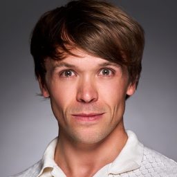 A man with brown hair wearing a white shirt, neutral expression, grey background.