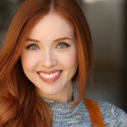 Smiling woman with long red hair, wearing a grey top and orange suspenders, neutral background.