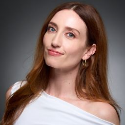 A woman with long brown hair, wearing a white top, poses against a grey background.