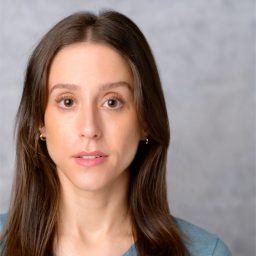 A woman with long brown hair and a blue top, against a grey background, looks forward.